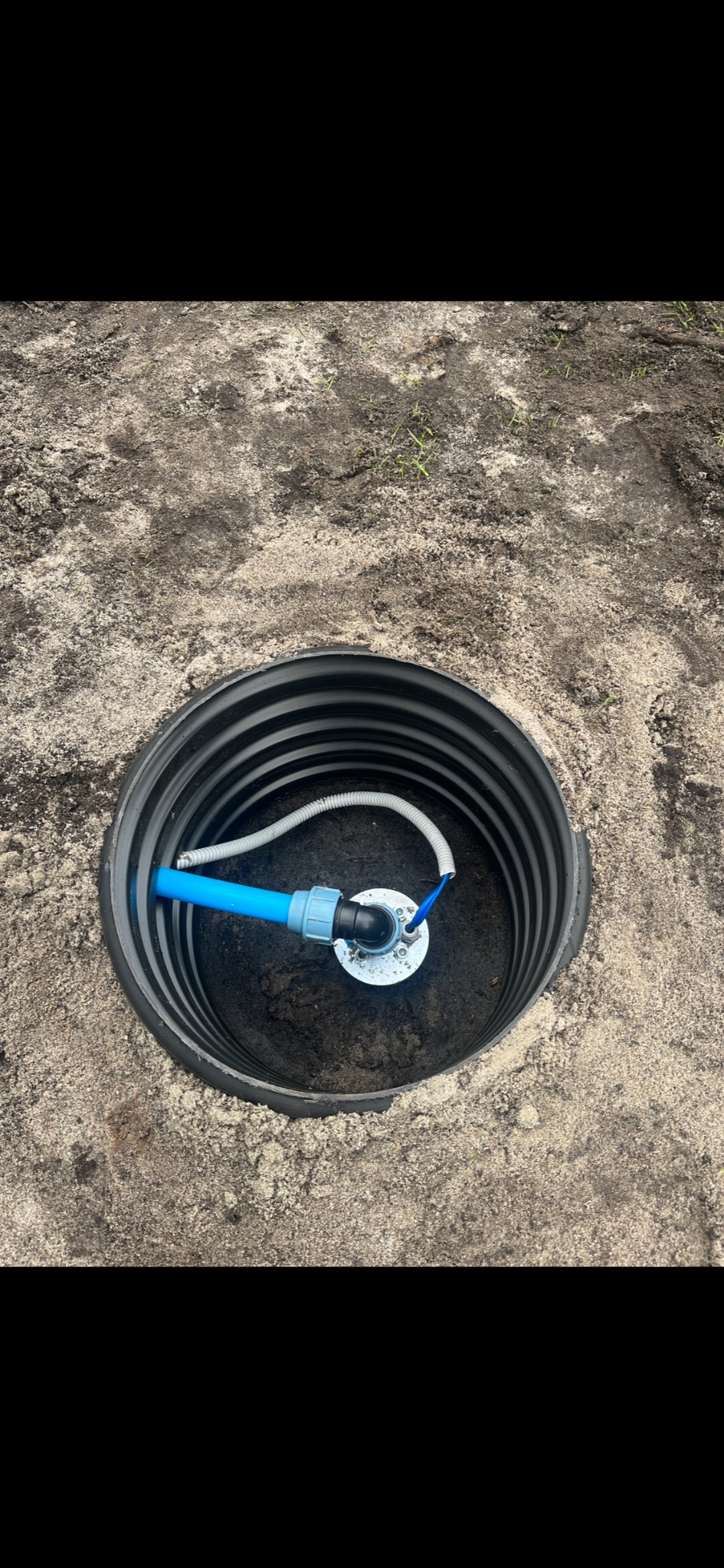 Czarna studzienka z niebieskim rurociągiem i szarym przewodem w piasku, widok z góry.