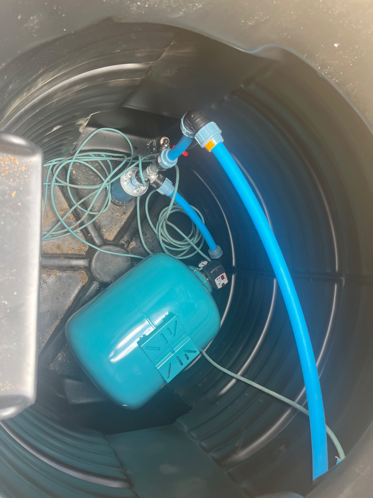 Wnętrze czarnego zbiornika z osprzętem hydroforowym: niebieski zbiornik ciśnieniowy, węże i przewody elektryczne.
