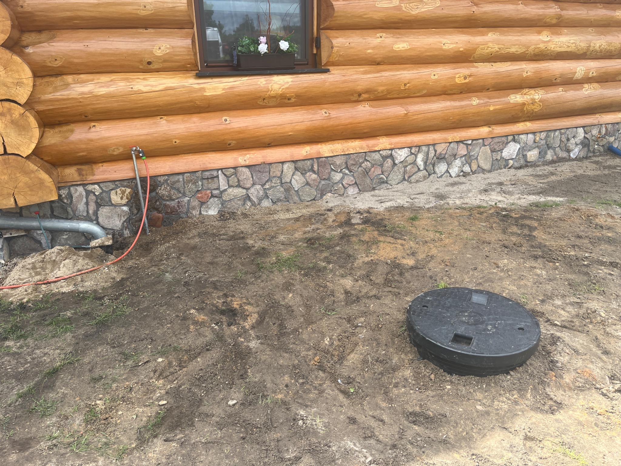 Studzienka rewizyjna z czarnym, okrągłym włazem na ziemi obok drewnianego domu z kamienną podmurówką i wystającym rurociągiem kanalizacyjnym.