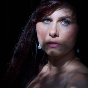 Portret kobiety z intensywnym spojrzeniem, oświetlona kontrastowym światłem i cieniem, z widocznymi kolczykami z perłami.