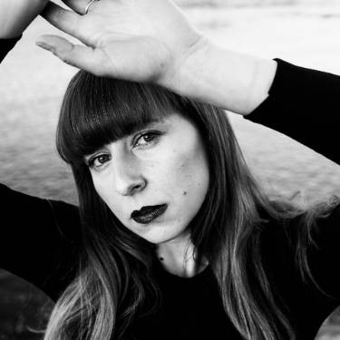 Portret kobiety z grzywką w czarno-białej tonacji, z ręką uniesioną nad czołem, ciemna szminka, długie włosy, w tle rozmyte tło.