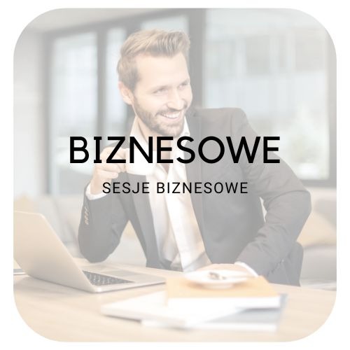 Uśmiechnięty mężczyzna w garniturze siedzi przy biurku z laptopem, książkami i filiżanką kawy, w tle rozmyte biuro.