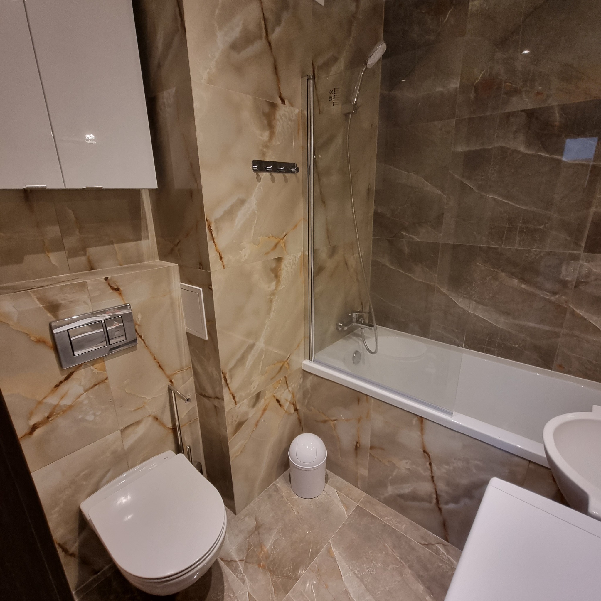 Wykończona łazienka z marmurowymi płytkami, białą armaturą i zabudową, widok na toaletę, wannę z parawanem i szafkę wiszącą.