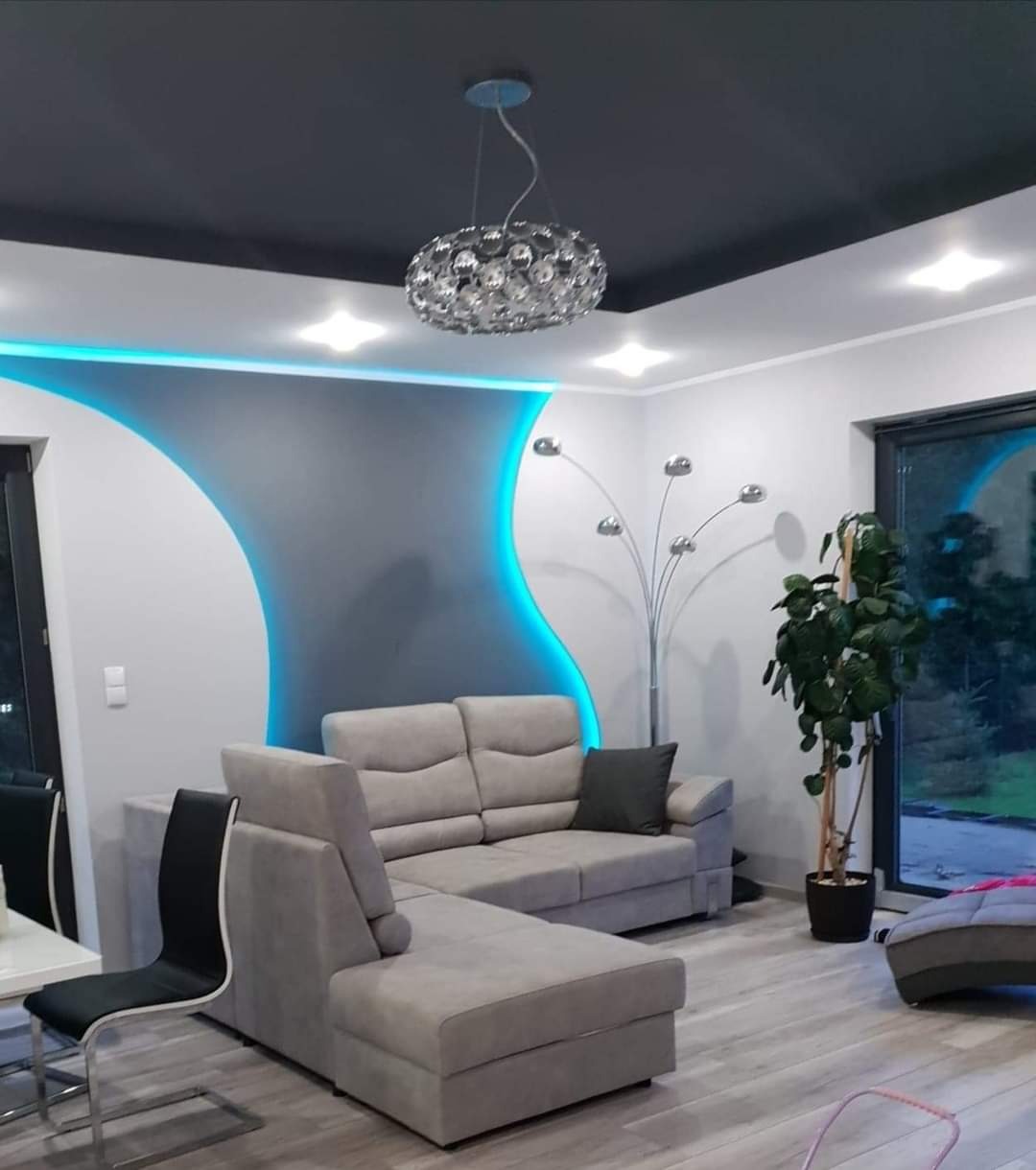 Nowoczesny salon z szarą sofą narożną, podświetlaną ścianą z niebieskim akcentem LED, czarnym sufitem podwieszanym i designerską lampą wiszącą.