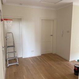 Przemysław Michalski - Pomalowane na biało wnętrze pokoju z drewnianą podłogą, drabina oparta o ścianę z zamontowanym koszem do koszykówki, puszki z farbą w rogu pomieszczenia.