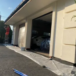 Mężczyzna maluje elewację domu jednorodzinnego na drabinie, zabezpieczone folią okna i podjazd, widoczny biały samoch&oacute;d w garażu.