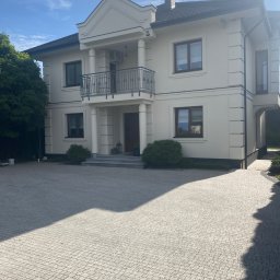 Przemysław Michalski - Elewacja domu jednorodzinnego w jasnych kolorach, z balkonem, kolumnami i oknami z ciemnymi ramami, widok z poziomu brukowanego podjazdu.