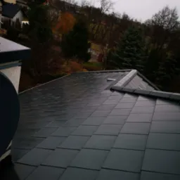 Świeżo ułożony, ciemnoszary dach z dachówki ceramicznej, widok z góry na tle zieleni drzew i zabudowań mieszkalnych w pochmurny dzień.