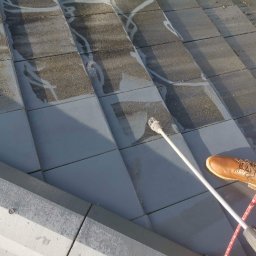 Przemysław Michalski - Czyszczenie dachówki ceramicznej z piasku i mchu za pomocą myjki ciśnieniowej, widoczny fragment dachu, dysza myjki, brązowy but pracownika i czerwony wąż myjki.