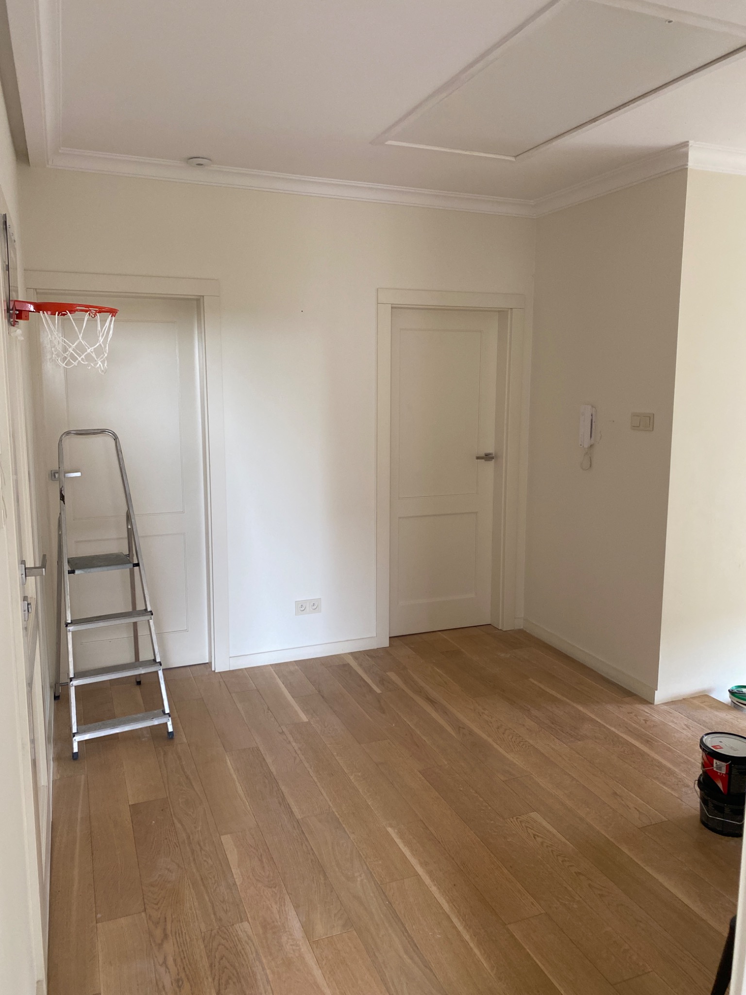 Pomalowane na biało wnętrze pokoju z drewnianą podłogą, drabina oparta o ścianę z zamontowanym koszem do koszykówki, puszki z farbą w rogu pomieszczenia.