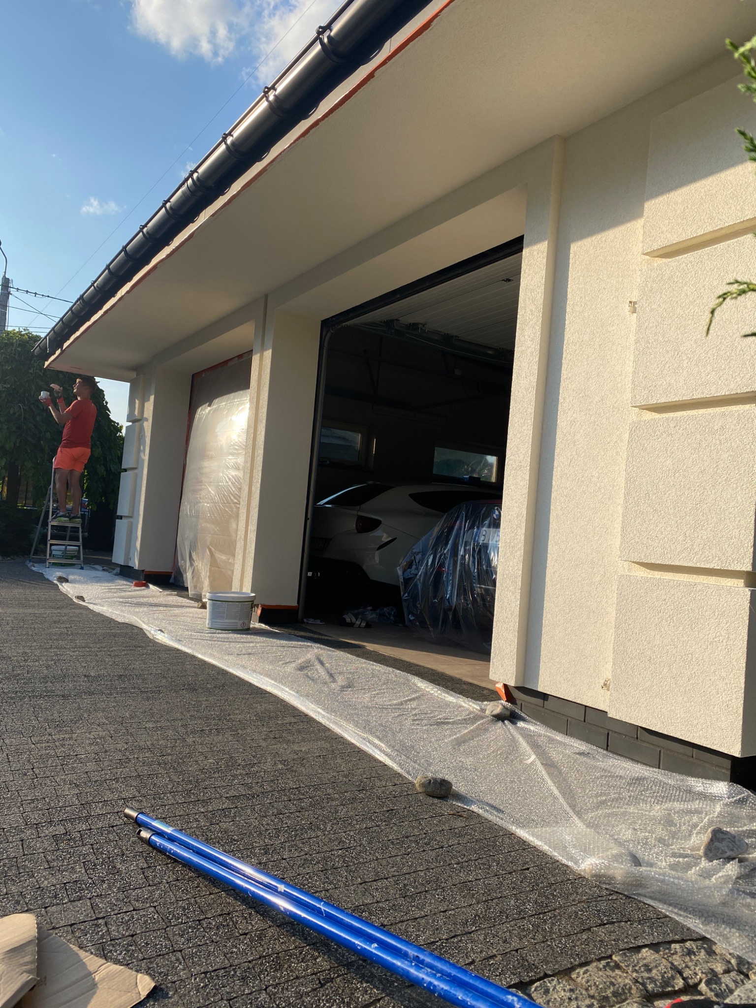 Mężczyzna maluje elewację domu jednorodzinnego na drabinie, zabezpieczone folią okna i podjazd, widoczny biały samochód w garażu.