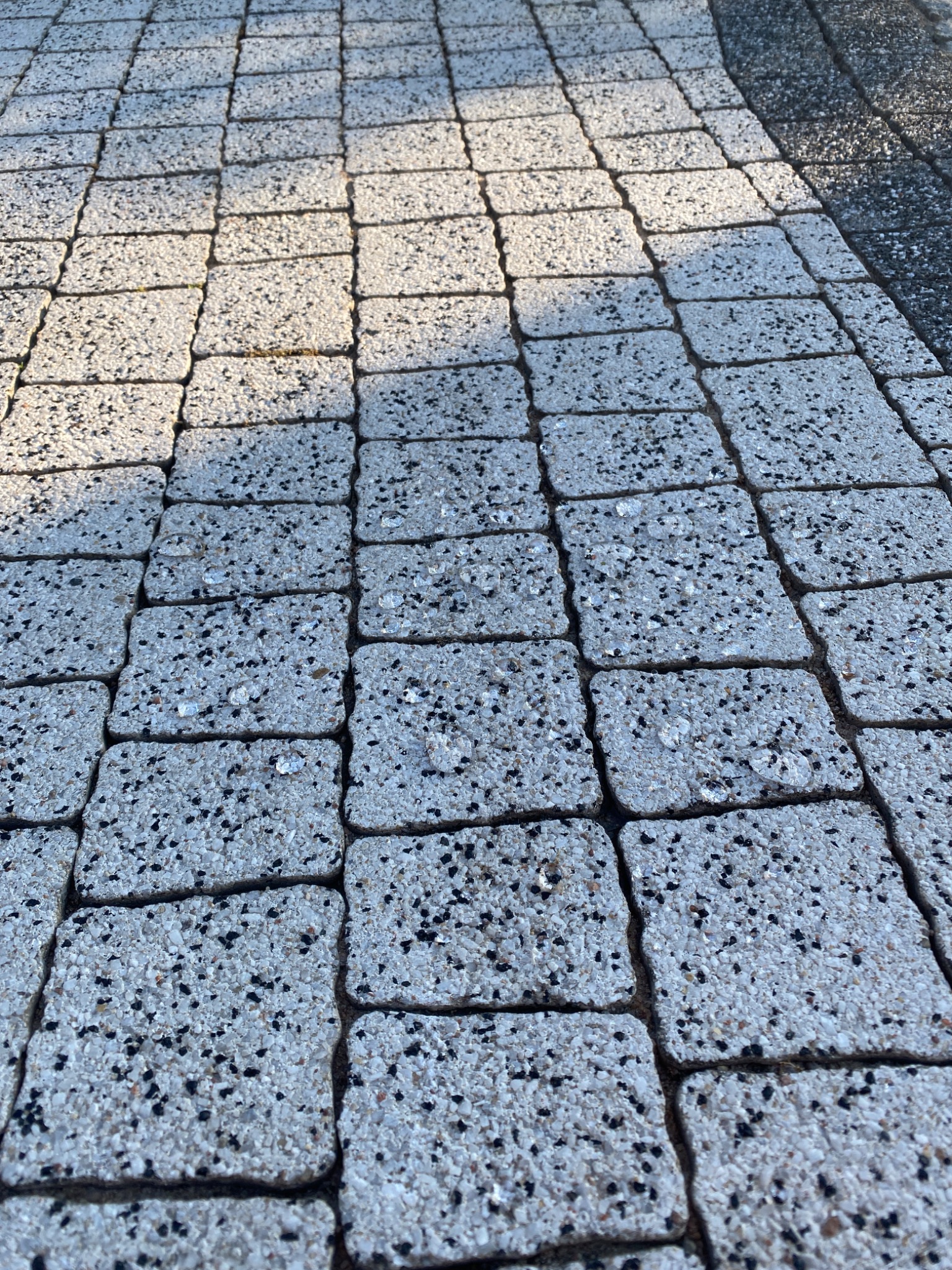 Szara kostka brukowa z czarnymi drobinkami, ułożona w regularny wzór, oświetlona nierównomiernie, tworząc cień na części powierzchni.