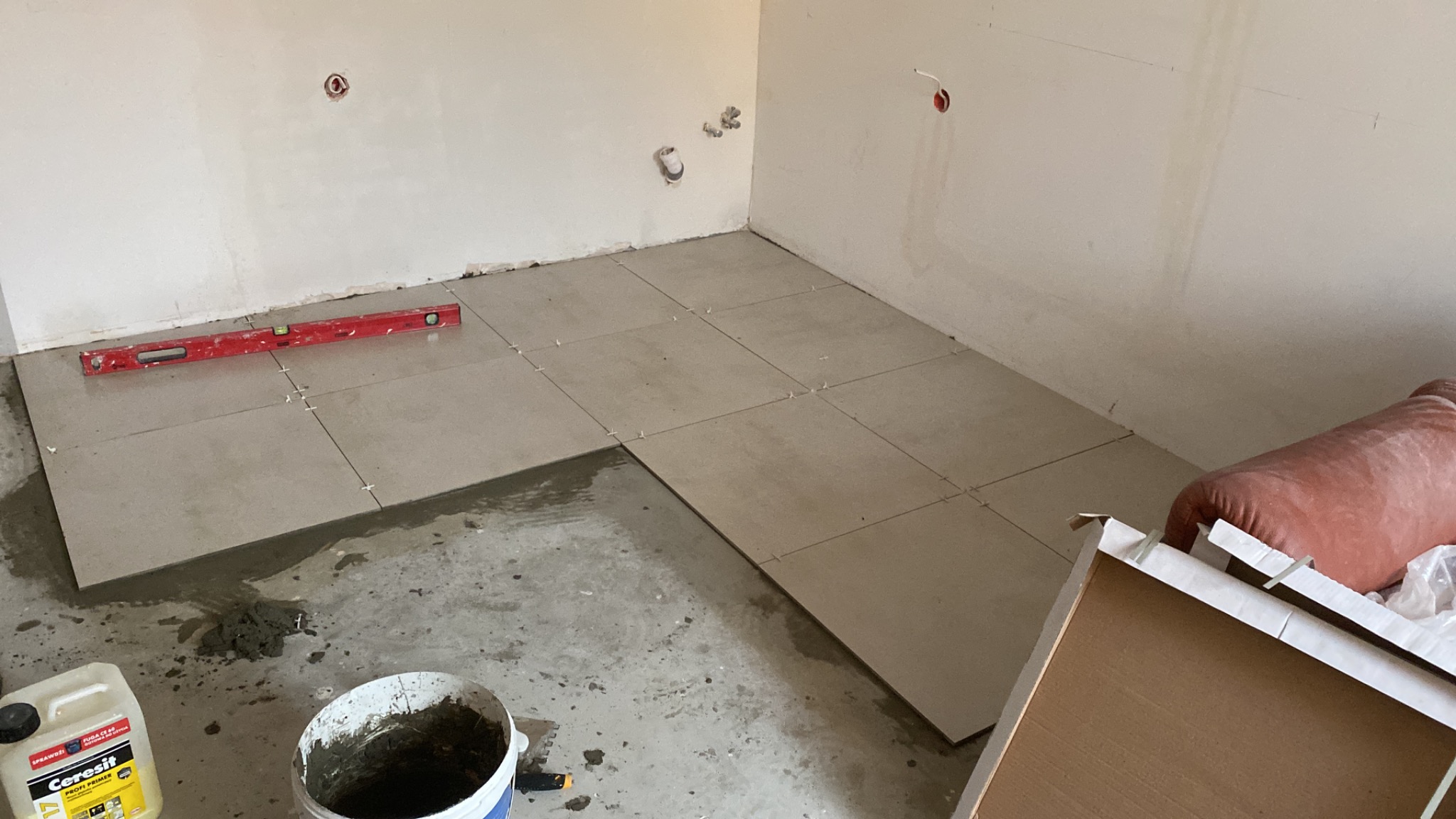 Wnętrze w trakcie układania szarych płytek podłogowych, widoczny poziomica, wiadro z zaprawą, krzyżyki dystansowe i grunt Cerisit
