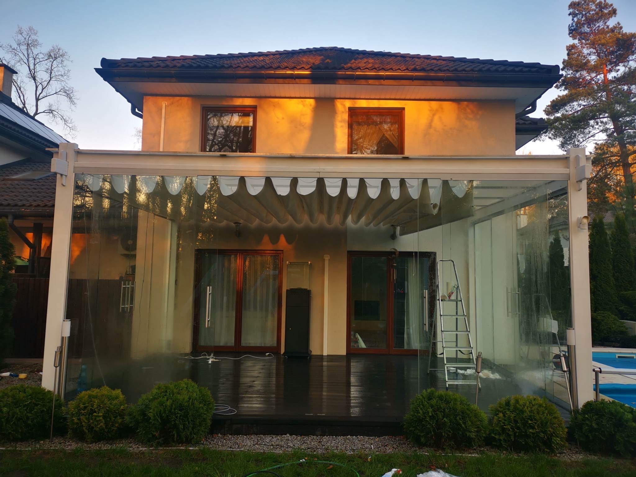 Zadaszony taras z przesuwnymi szklanymi ścianami, drewnianą podłogą i zewnętrznym oświetleniem, widoczny z zewnątrz, z drabiną opartą o jedną ze ścian, tuż po umyciu.