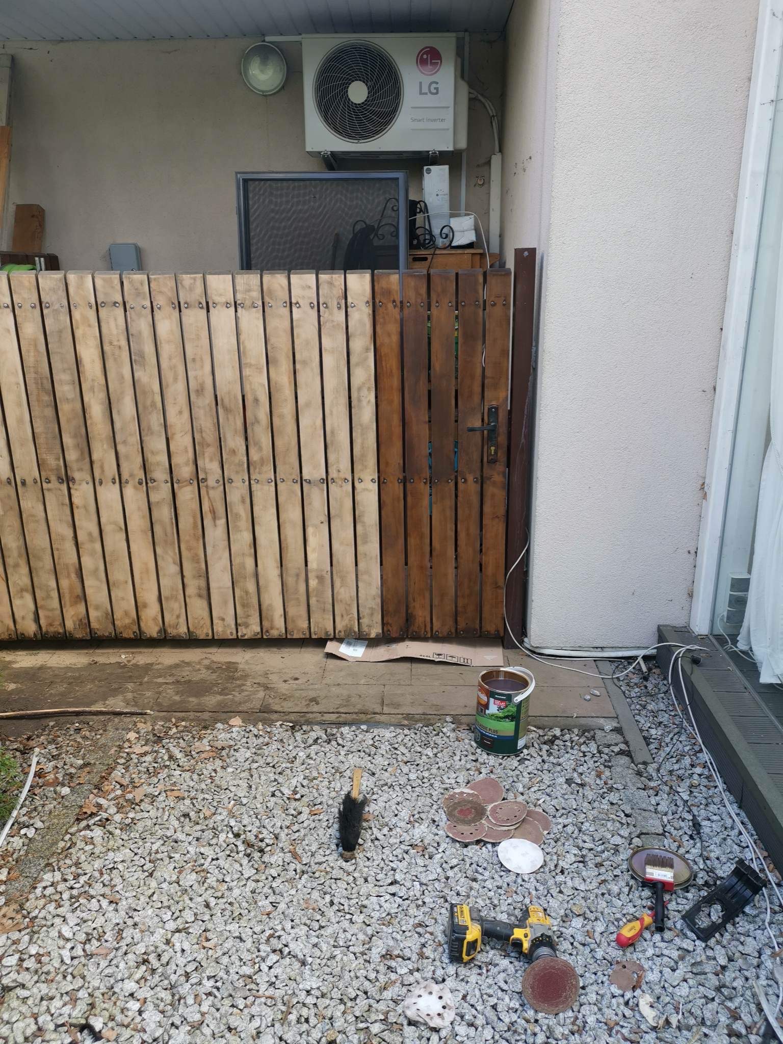 Drewniana brama, częściowo pomalowana na brązowo, widoczna różnica przed i po malowaniu, obok puszka farby, szlifierka, pędzle i inne narzędzia na żwirowym podłożu.