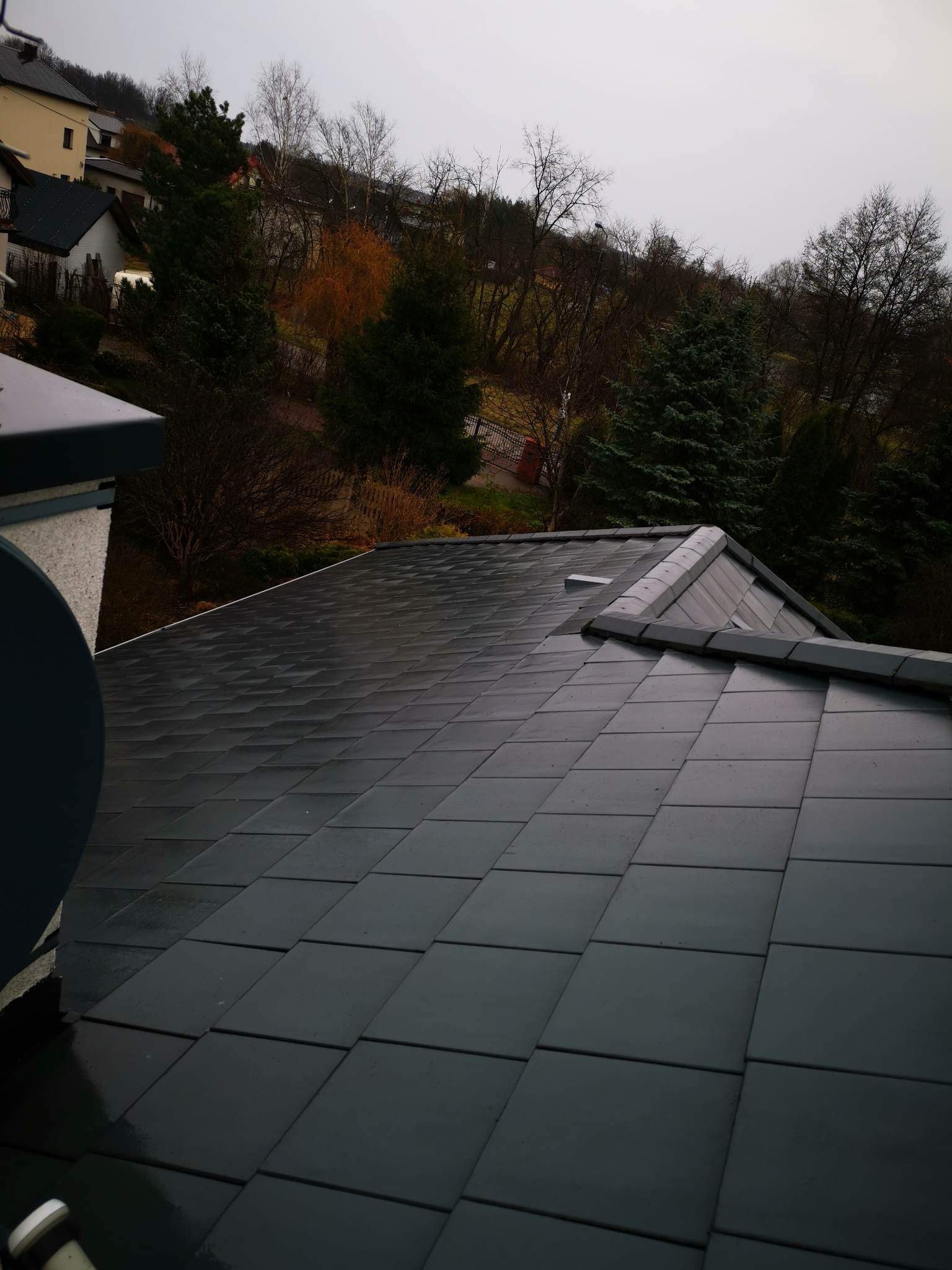 Świeżo ułożony, ciemnoszary dach z dachówki ceramicznej, widok z góry na tle zieleni drzew i zabudowań mieszkalnych w pochmurny dzień.