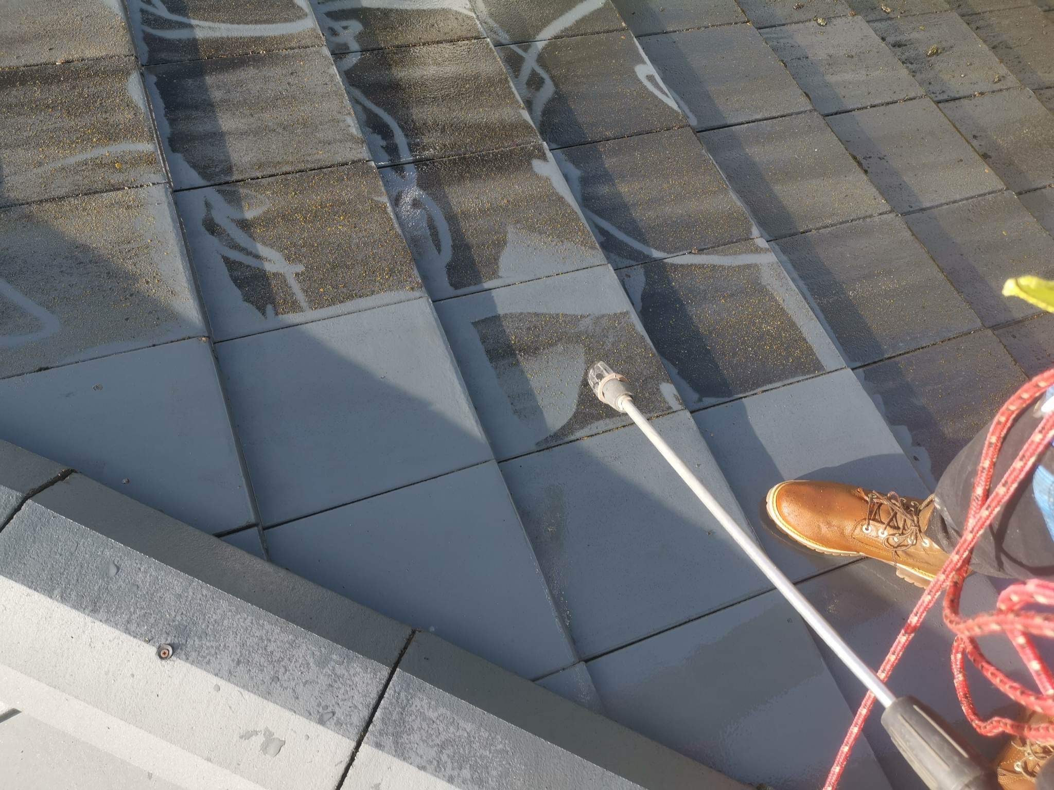 Czyszczenie dachówki ceramicznej z piasku i mchu za pomocą myjki ciśnieniowej, widoczny fragment dachu, dysza myjki, brązowy but pracownika i czerwony wąż myjki.