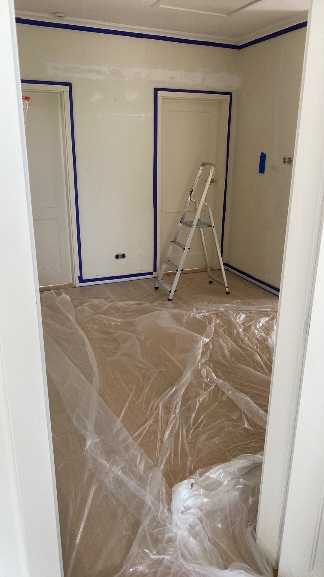 Pokój przygotowany do malowania: podłoga zabezpieczona folią, ściany i futryny oklejone niebieską taśmą malarską, aluminiowa drabina rozstawiona przy ścianie.