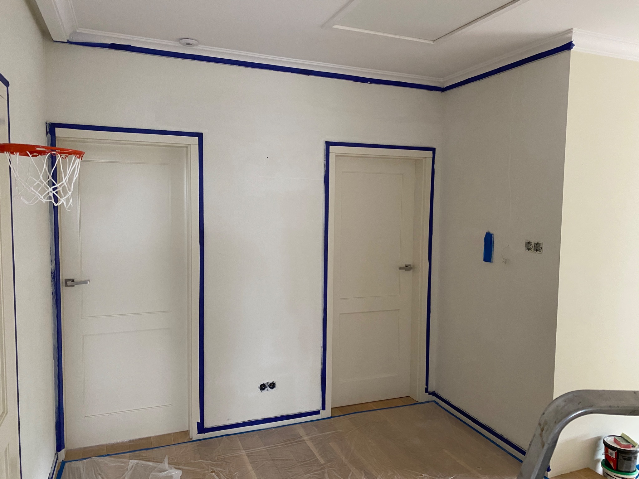 Pokój w trakcie zabezpieczania przed malowaniem: białe ściany i drzwi oklejone niebieską taśmą malarską, podłoga przykryta folią ochronną, widoczny kosz do koszykówki i drabina z farbami.
