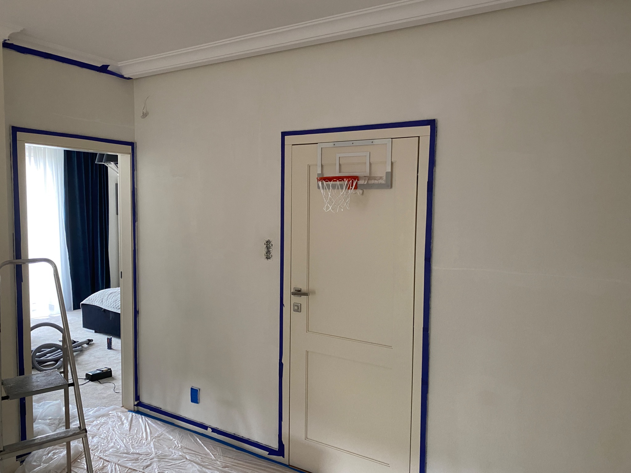 Pokój przygotowany do malowania: ściany zabezpieczone taśmą malarską, podłoga przykryta folią ochronną, drabina i odkurzacz widoczne w tle, mały kosz do koszykówki zawieszony na drzwiach.