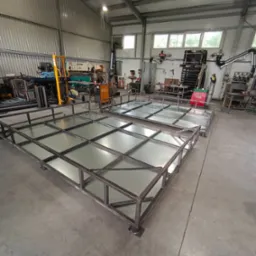 Dwa metalowe ruszty z kwadratowymi polami, prawdopodobnie kraty pomostowe, w hali produkcyjnej z widocznym wyposażeniem warsztatowym w tle.