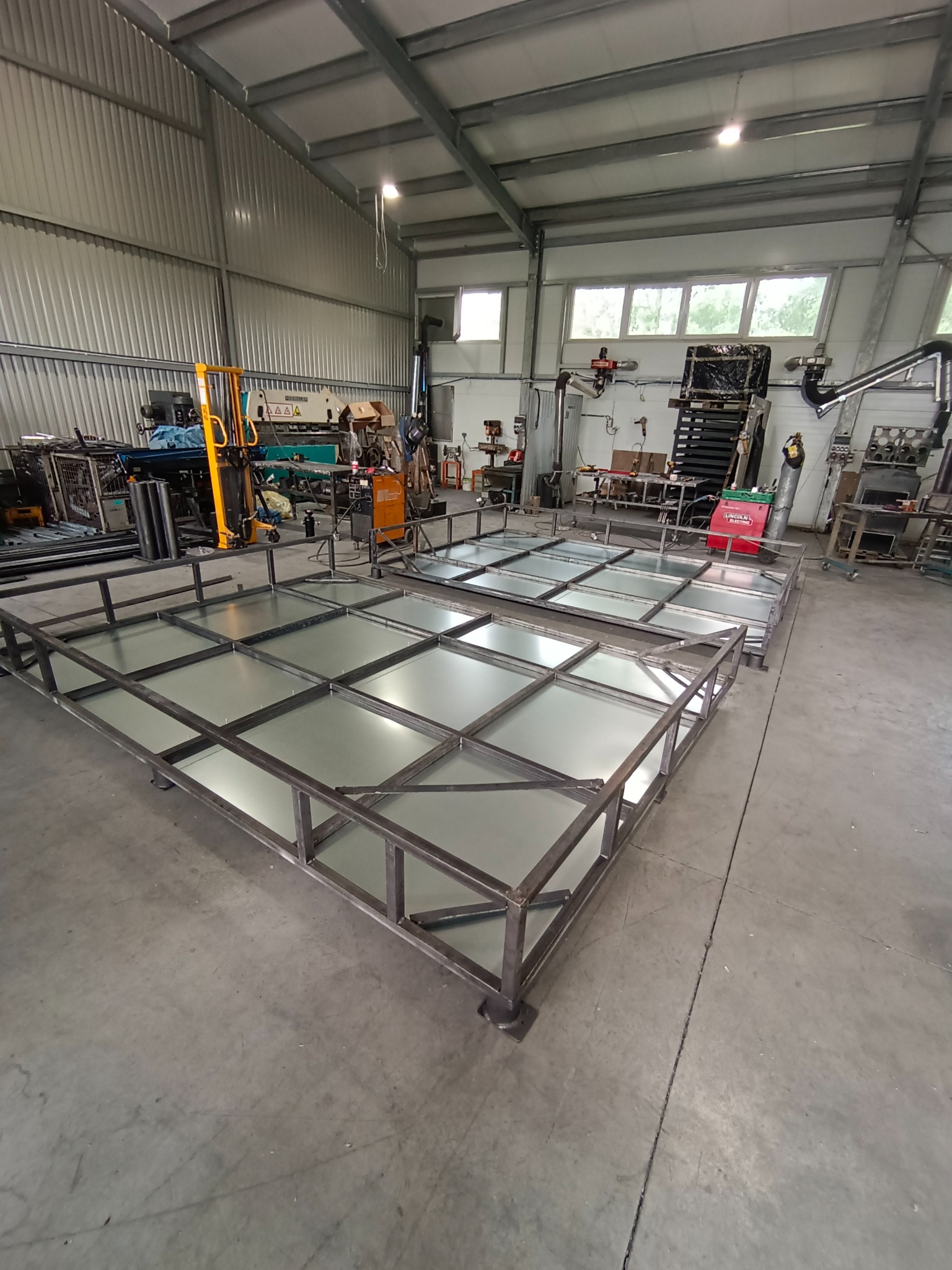 Dwa metalowe ruszty z kwadratowymi polami, prawdopodobnie kraty pomostowe, w hali produkcyjnej z widocznym wyposażeniem warsztatowym w tle.