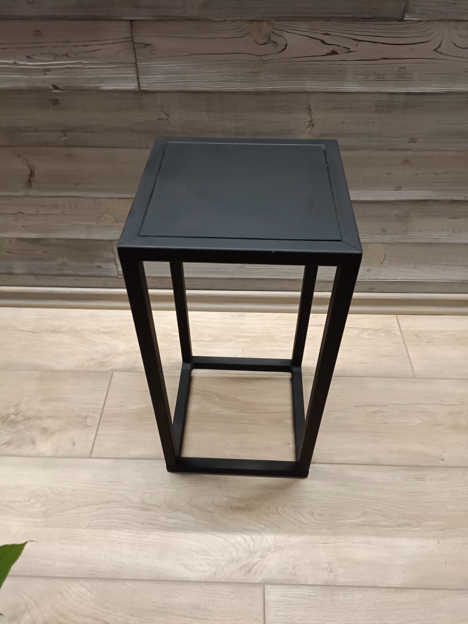 Czarny, metalowy stolik kawowy o minimalistycznym designie, z kwadratowym blatem i ramą, na tle drewnianej ściany i podłogi z paneli.