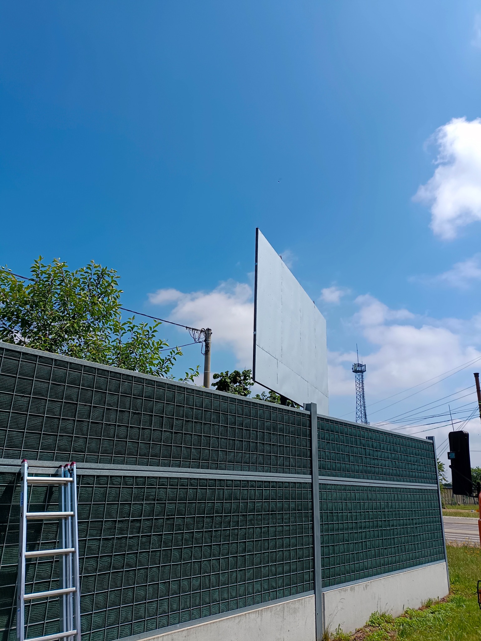 Długi, metalowy ekran akustyczny z siatkową strukturą, wsparty na betonowej podstawie, z aluminiową drabiną opartą o jego bok, na tle błękitnego nieba z chmurami.