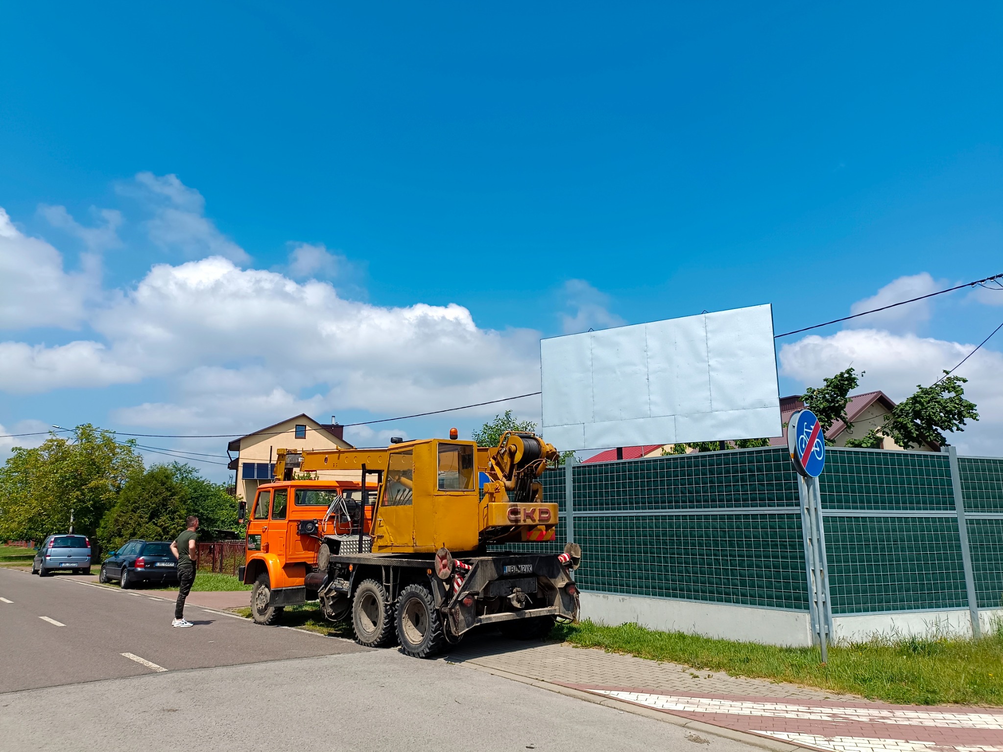 Pomarańczowo-żółty dźwig samochodowy CKD zaparkowany na poboczu drogi obok zielonego ekranu akustycznego, z mężczyzną obserwującym pracę w tle.