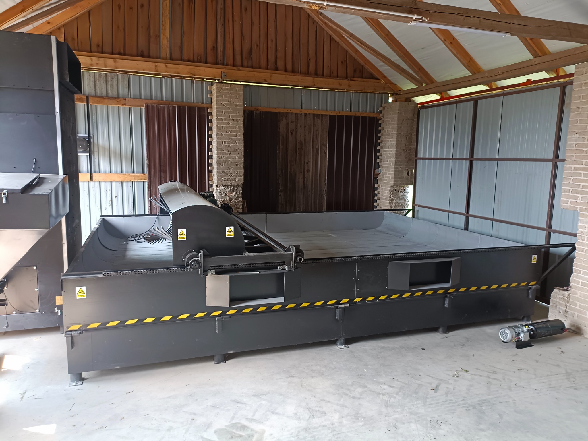 Duże, czarne urządzenie do obróbki metali z żółtymi pasami ostrzegawczymi, umieszczone w hali z ceglanymi filarami i drewnianą konstrukcją dachu.