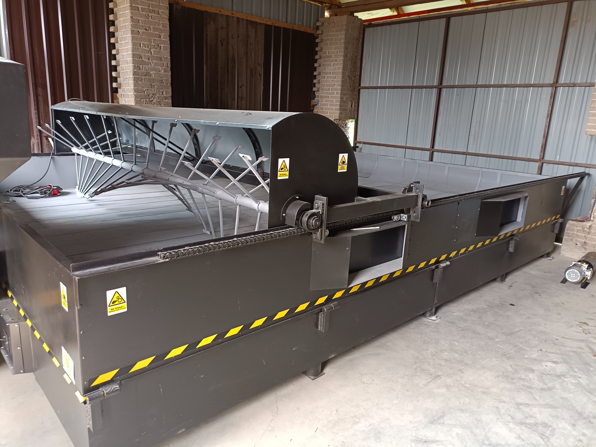 Metalowa konstrukcja z elementami ruchomymi, prawdopodobnie część maszyny przemysłowej, z żółto-czarnymi pasami ostrzegawczymi, umieszczona w hali z ceglanym filarem.