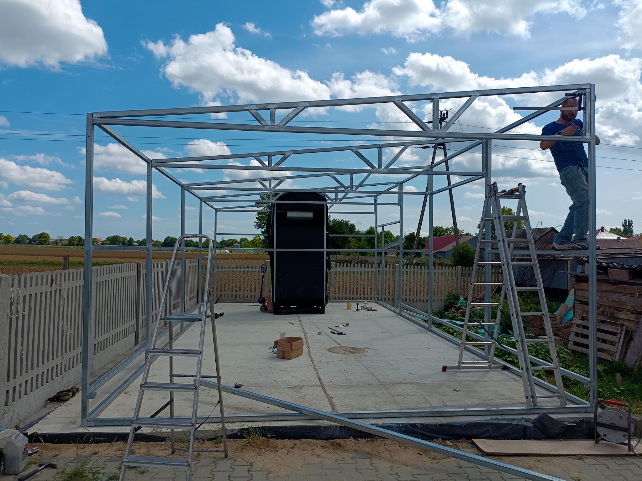 Konstrukcja szkieletowa z metalowych profili w trakcie montażu na betonowej wylewce, pracownik na drabinie mocuje górne elementy, w tle ogrodzenie i błękitne niebo.