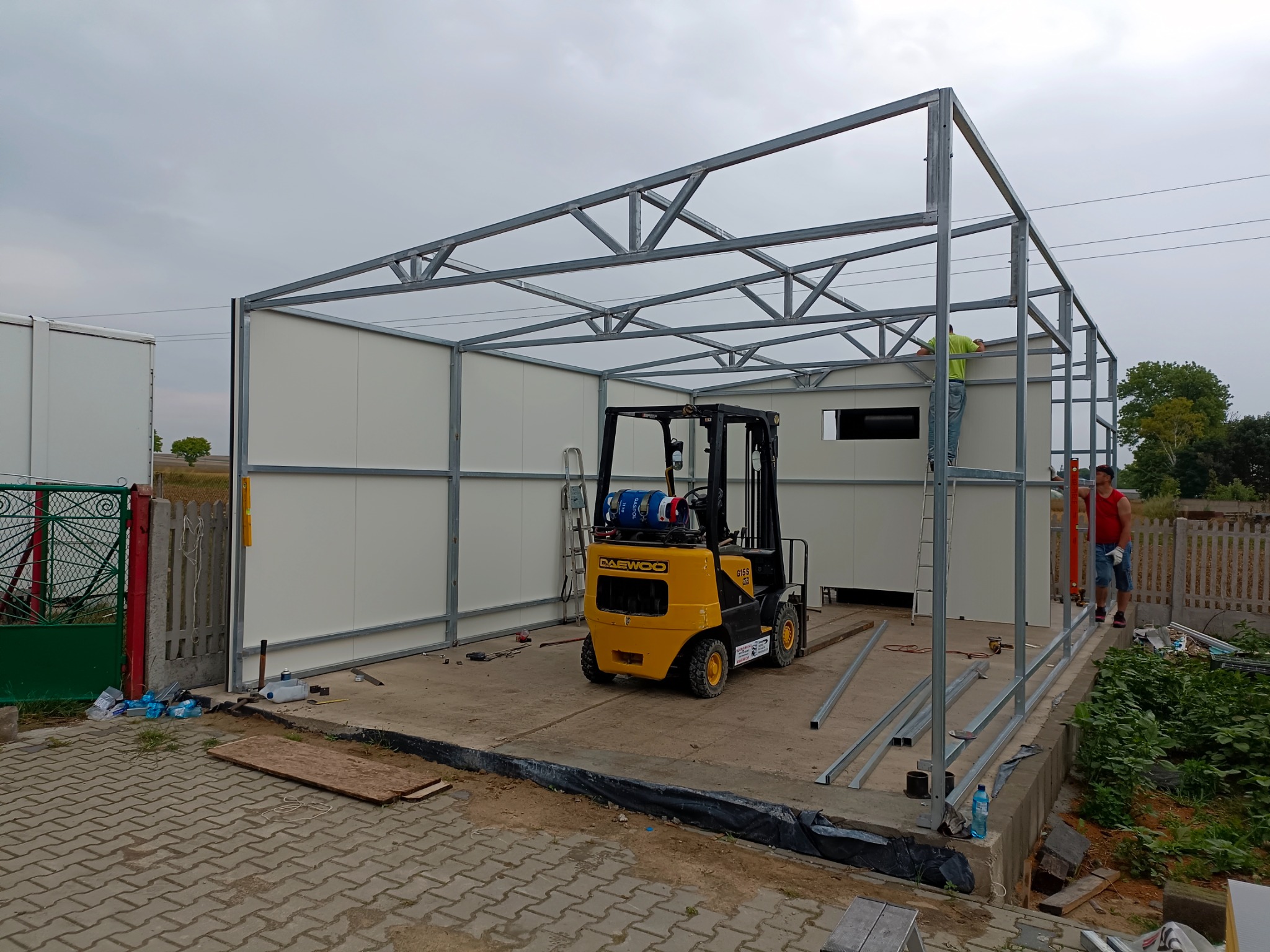 Żółty wózek widłowy Daewoo na tle szkieletu budowli z metalowych profili i białych paneli, z pracownikami w trakcie montażu.
