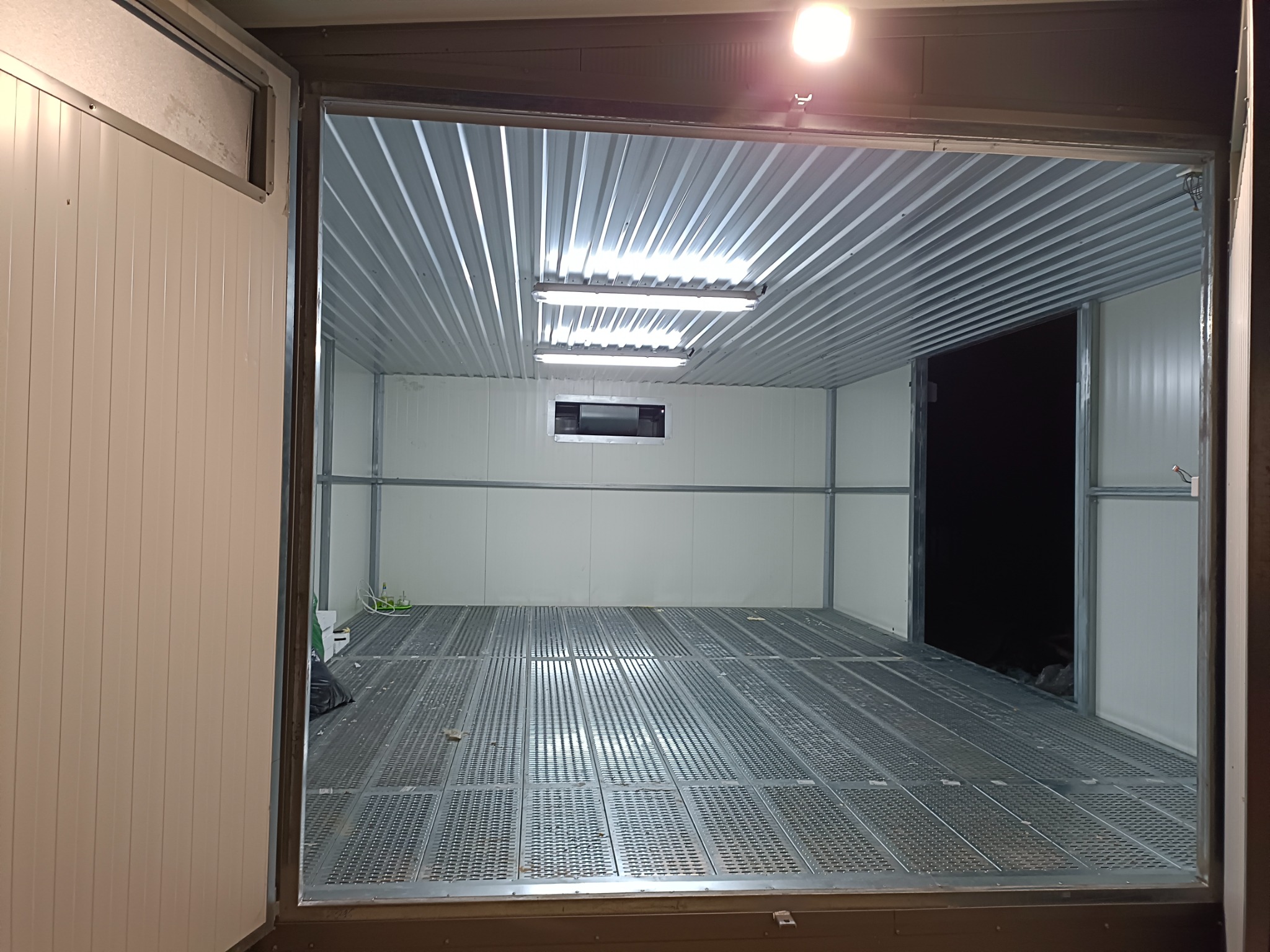 Wnętrze metalowej konstrukcji z perforowaną podłogą, oświetlone lampami jarzeniowymi i punktowym światłem, widoczne przez otwarte metalowe wrota.