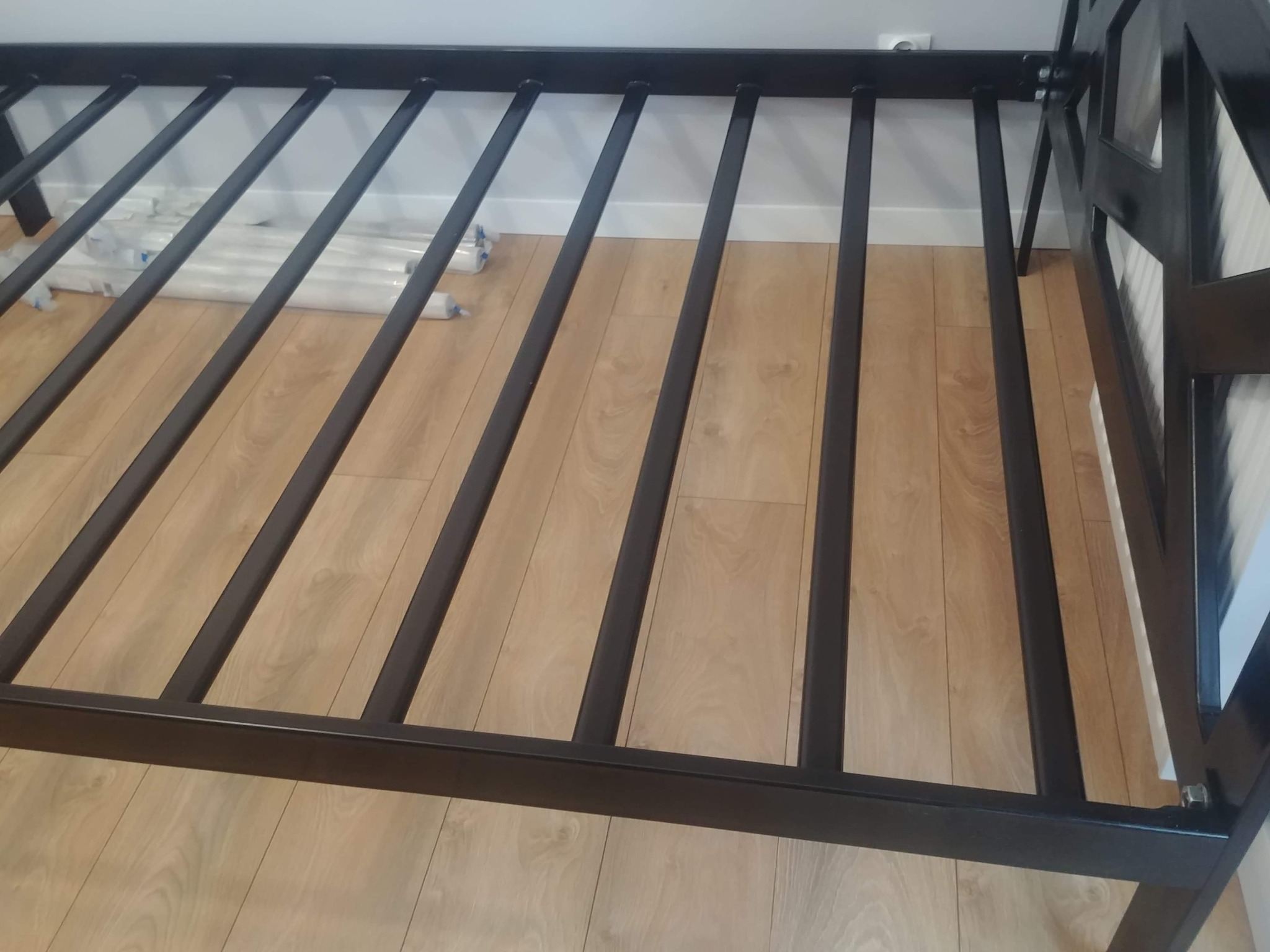 Metalowa konstrukcja ramy łóżka z poprzecznymi belkami, widoczne elementy montażowe i opakowania po elementach na podłodze.