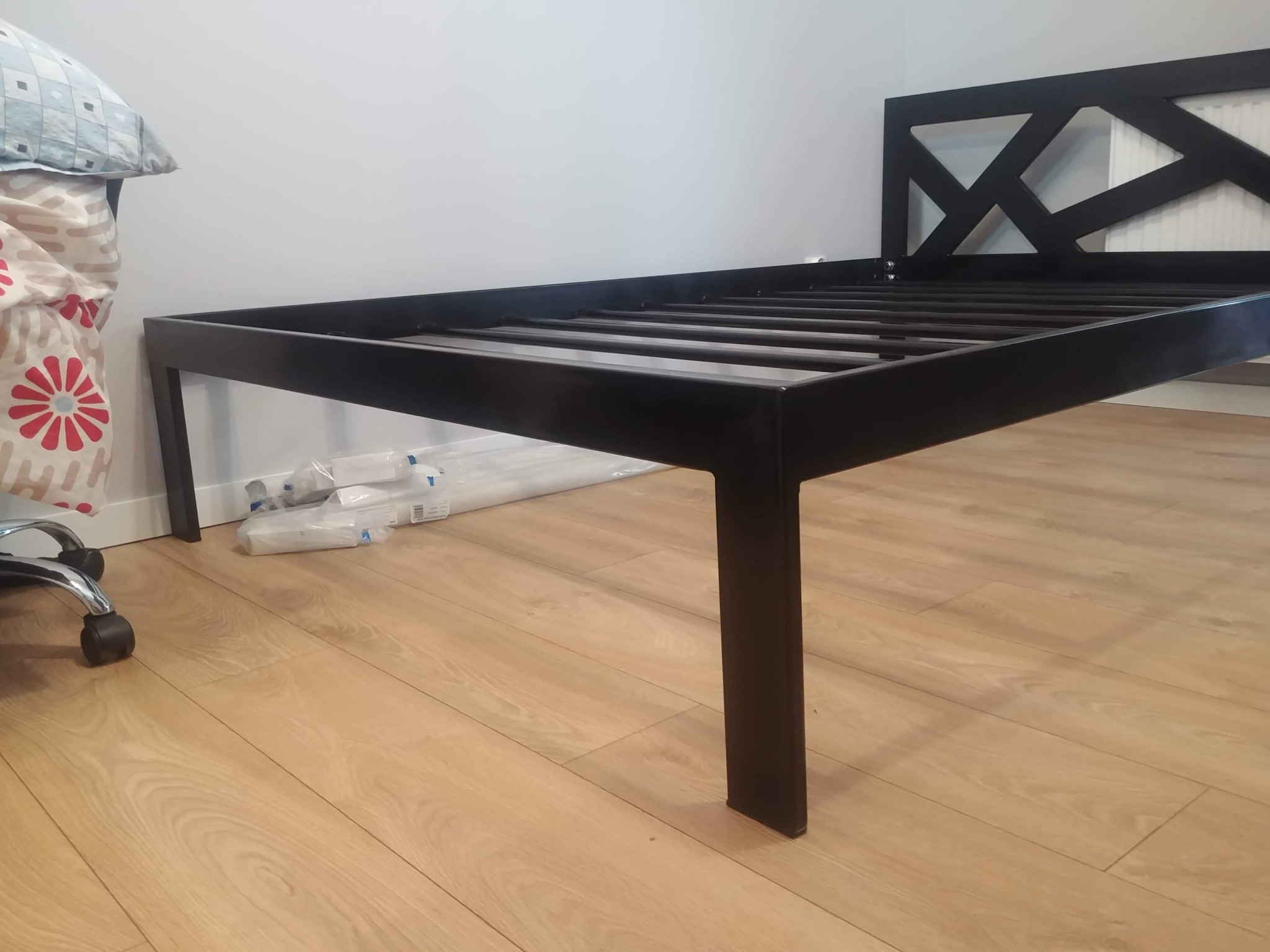 Czarna, metalowa rama łóżka z zagłówkiem o geometrycznym wzorze, ustawiona na parkiecie, z widocznymi elementami montażowymi i opakowaniami w tle.