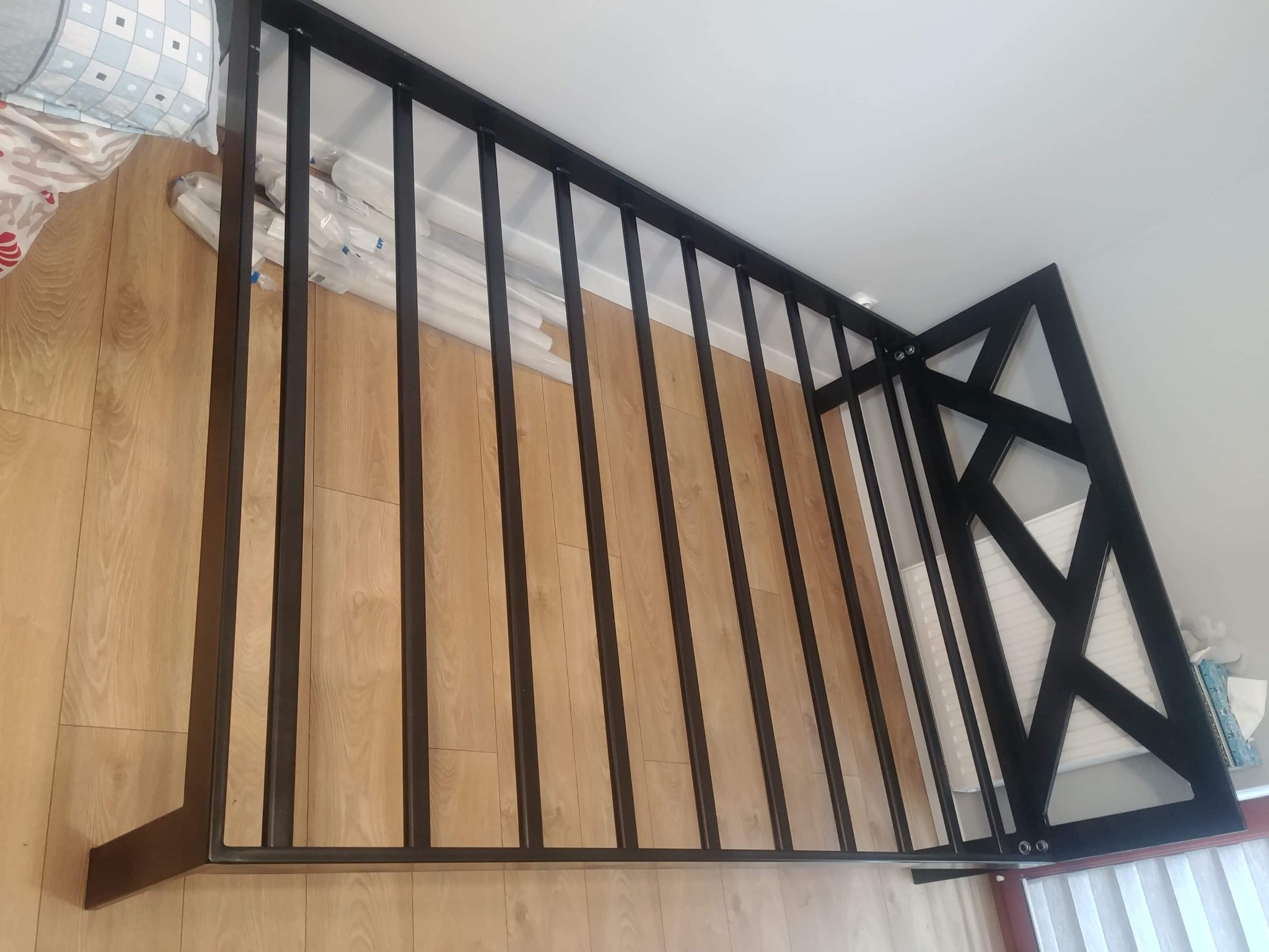 Czarna, metalowa balustrada schodowa z geometrycznym wzorem, częściowo zamontowana, leżąca na podłodze z paneli, widok z góry.