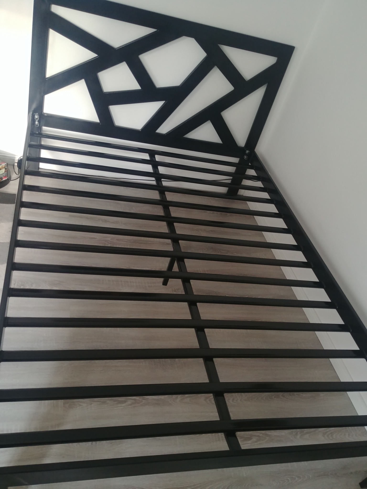 Metalowa rama łóżka z geometrycznym zagłówkiem w czarnym kolorze, widoczne drewniane listwy pod materac, minimalistyczny design.