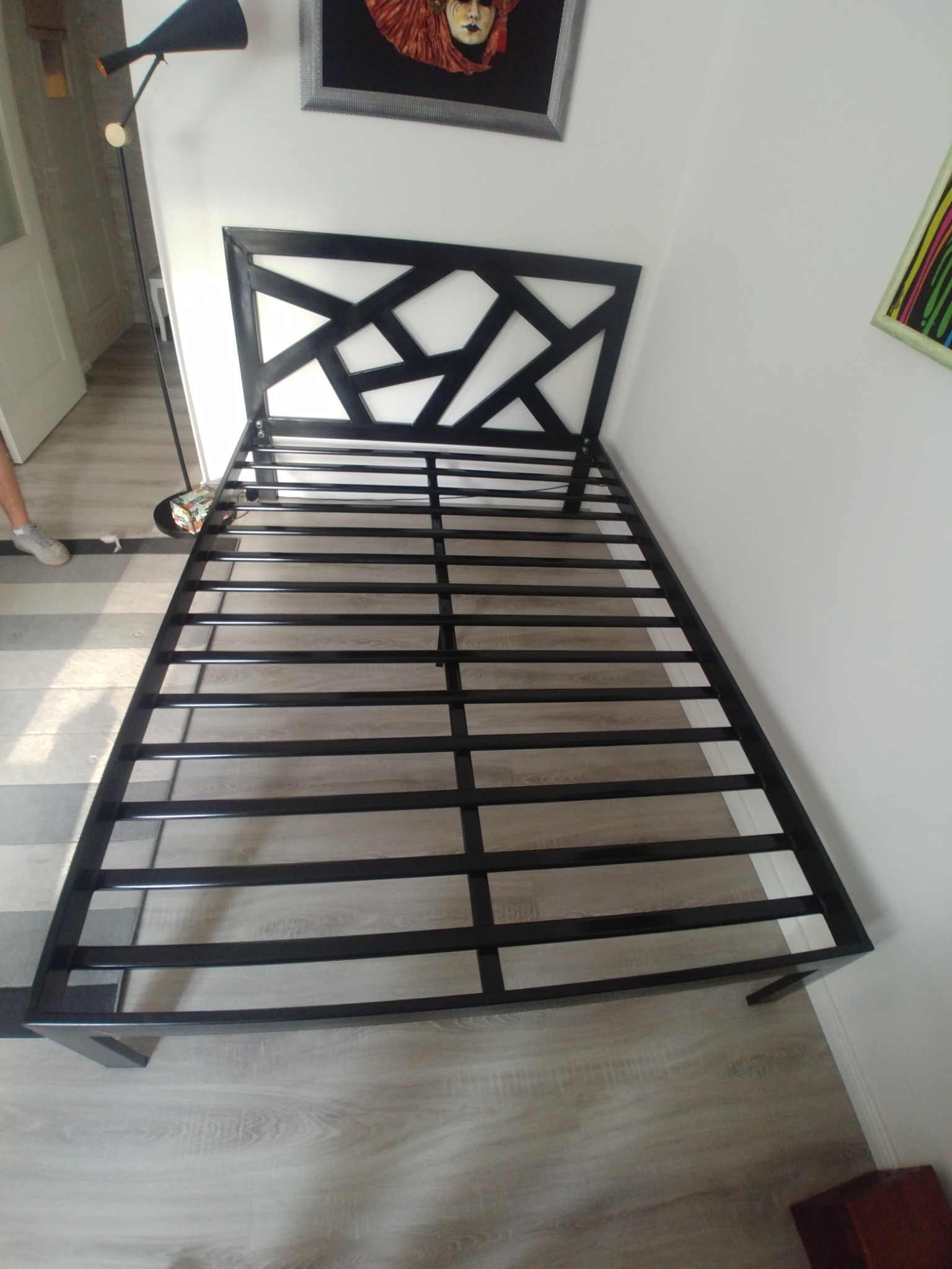 Metalowa rama łóżka z geometrycznym zagłówkiem, czarna konstrukcja na drewnopodobnej podłodze, widok z góry.