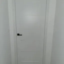 Zamknięte białe drzwi bezprzylgowe z czarną klamką, z czterema poziomymi wgłębieniami, osadzone w białej futrynie w białej ścianie, widok z dołu.