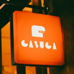 Pomarańczowy, podświetlany szyld kwadratowy z białym logo i napisem 'CABUCA' zawieszony na ulicy.