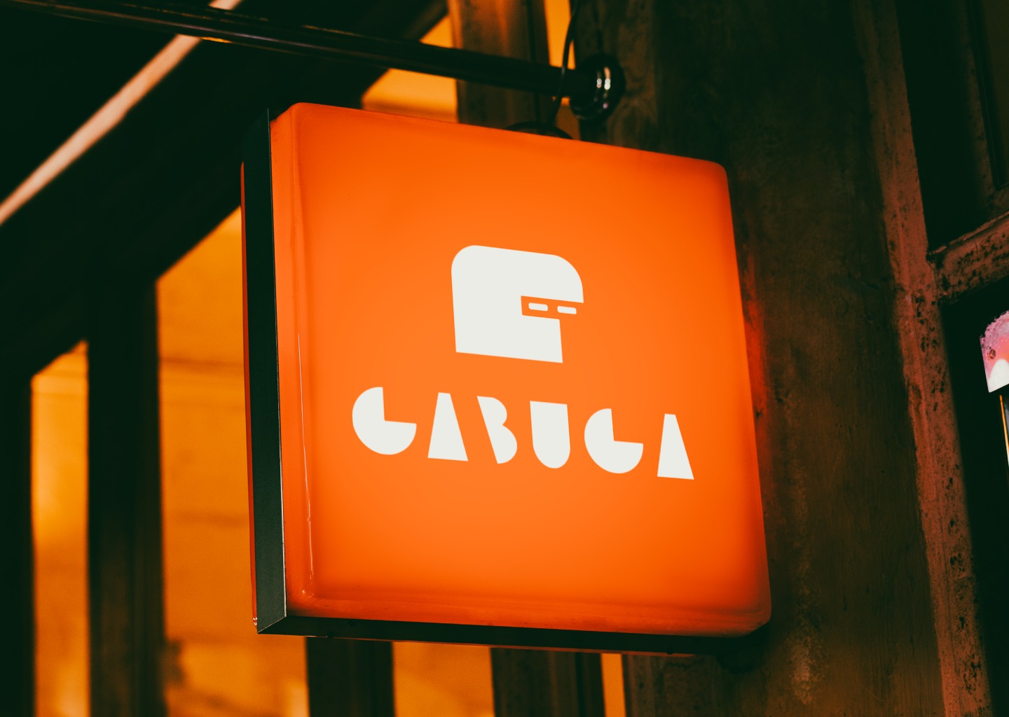 Pomarańczowy, podświetlany szyld kwadratowy z białym logo i napisem 'CABUCA' zawieszony na ulicy.