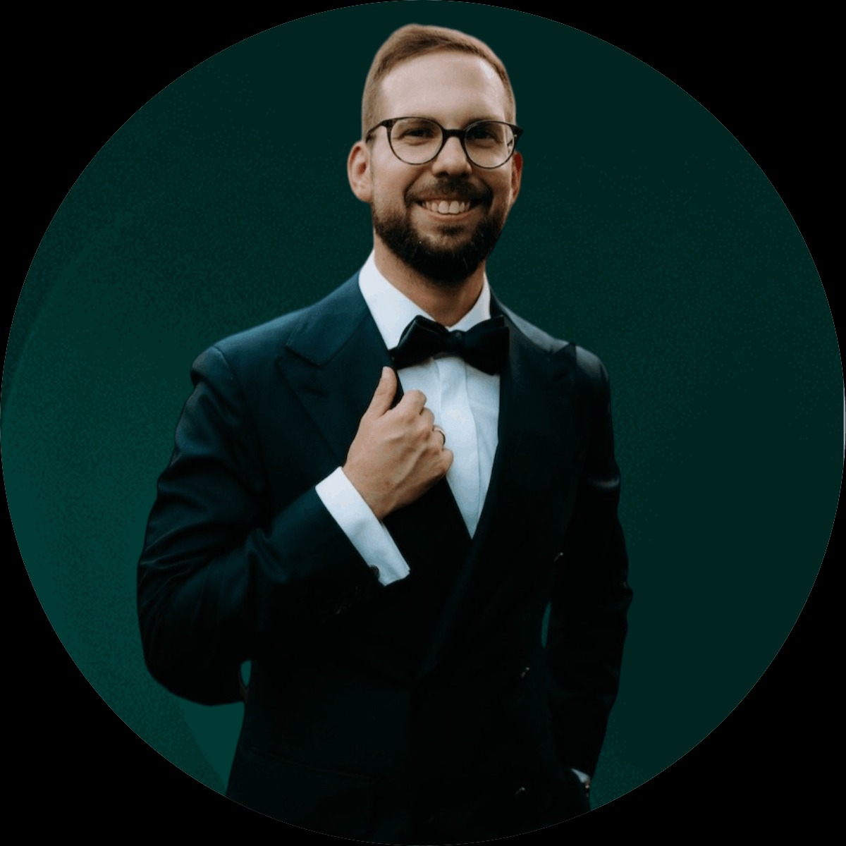 Portret uśmiechniętego mężczyzny w okularach i eleganckim smokingu z muszką na ciemnozielonym tle.