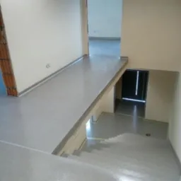 Jasnoszara, błyszcząca podłoga schodów i korytarza w trakcie remontu z widocznymi ścianami w odcieniach beżu i fragmentem ściany z cegłami.