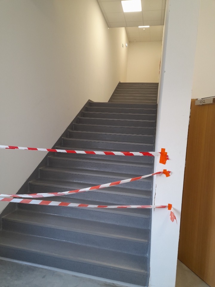 Schody w budynku biurowym, zabezpieczone taśmą ostrzegawczą, świeżo po malowaniu szarą farbą.