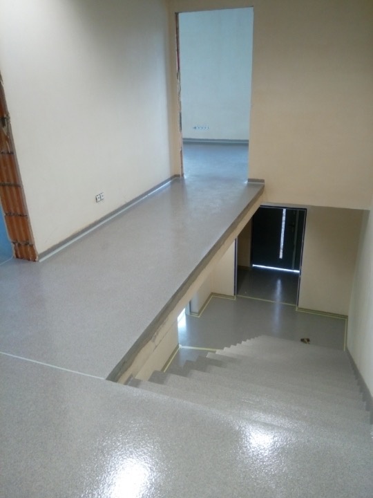 Jasnoszara, błyszcząca podłoga schodów i korytarza w trakcie remontu z widocznymi ścianami w odcieniach beżu i fragmentem ściany z cegłami.