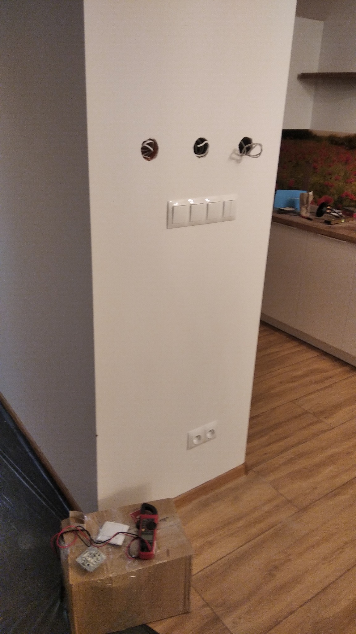 Biała ściana z trzema otworami pod instalację elektryczną, zamontowanym poczwórnym włącznikiem światła i gniazdkiem, na podłodze karton z miernikiem i przewodami, w tle fragment kuchni.