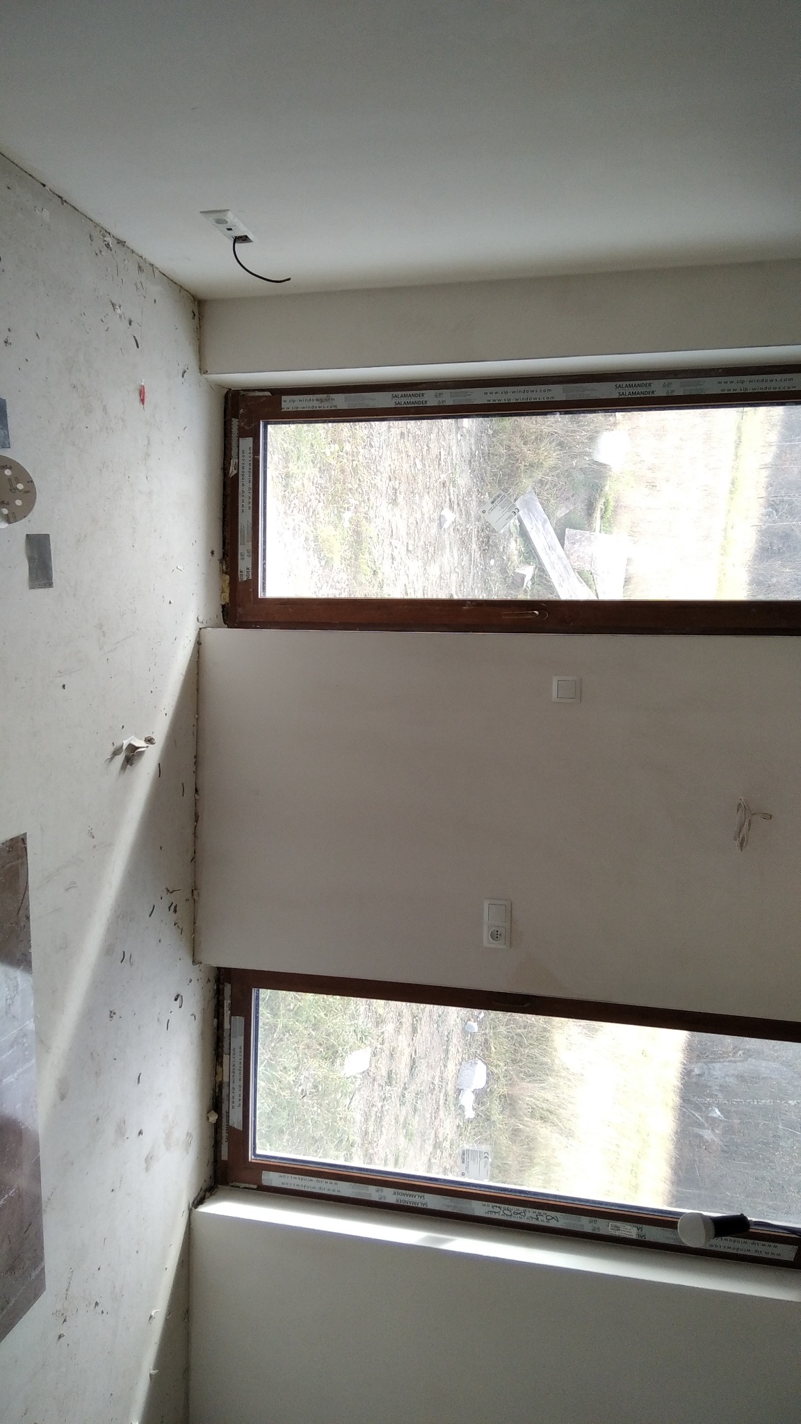 Wnętrze w trakcie remontu, widoczne dwa okna z brązowymi ramami, ściany częściowo otynkowane, gniazdko elektryczne wystające z sufitu z podłączonym przewodem oraz dwa gniazdka i włączniki na ścianie.