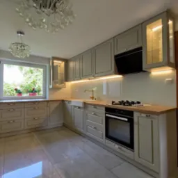 Jasnoszara kuchnia z drewnianym blatem, białym zlewem farmerskim i szklanymi szafkami z oświetleniem, widziana z perspektywy szerokokątnej, z dekoracyjnym żyrandolem w tle.