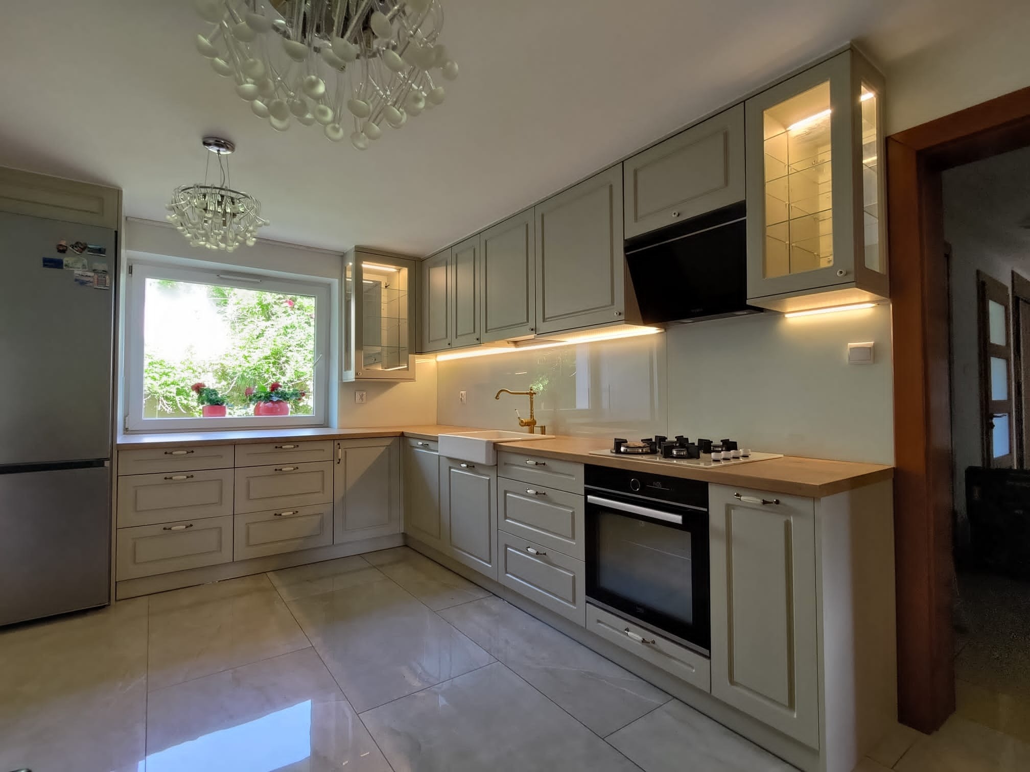 Jasnoszara kuchnia z drewnianym blatem, białym zlewem farmerskim i szklanymi szafkami z oświetleniem, widziana z perspektywy szerokokątnej, z dekoracyjnym żyrandolem w tle.